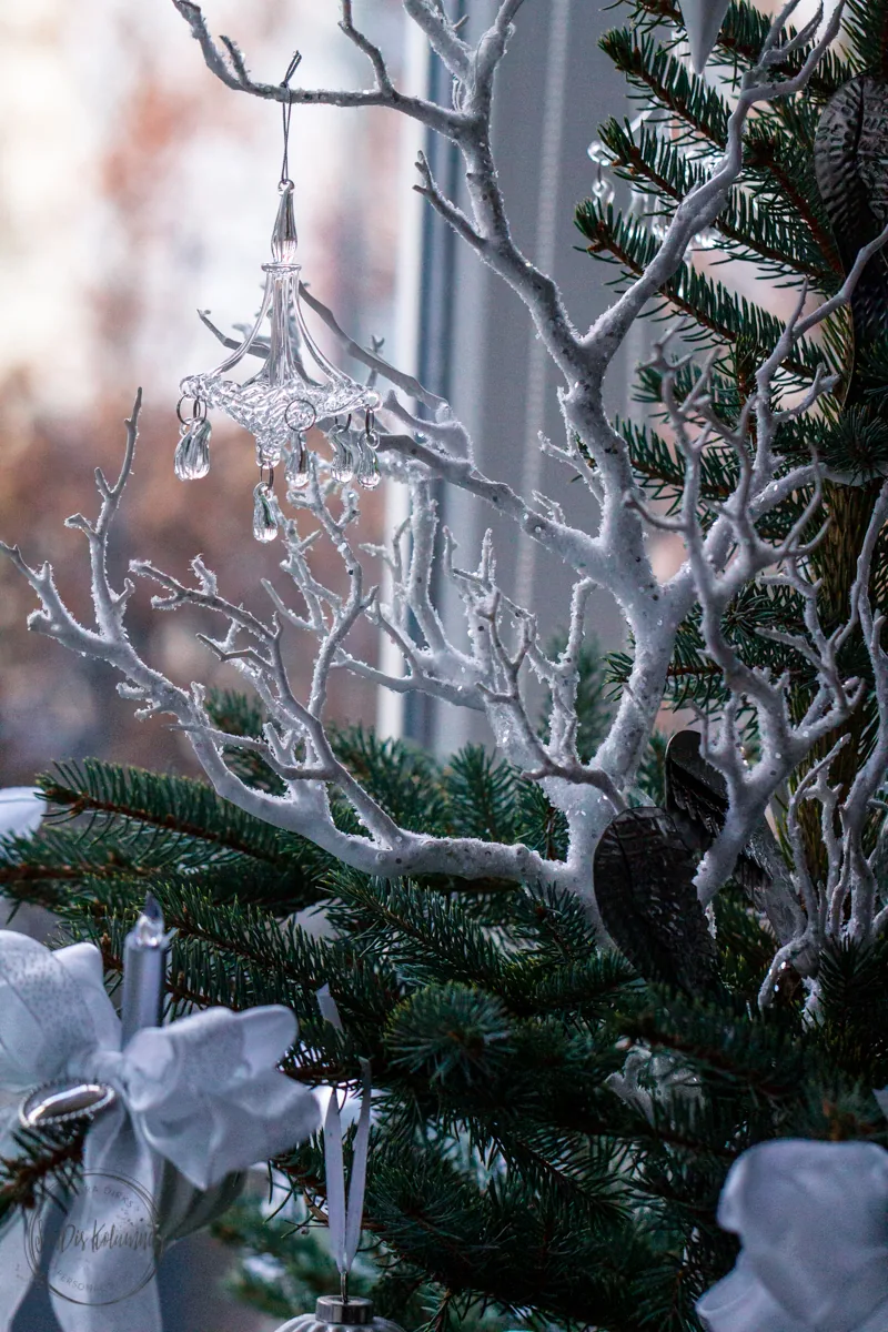 Gläserner Kronleuchter-Anhänger zwischen weiß glitzernden Zweigen und Tannengrün, dahinter das helle Fenster