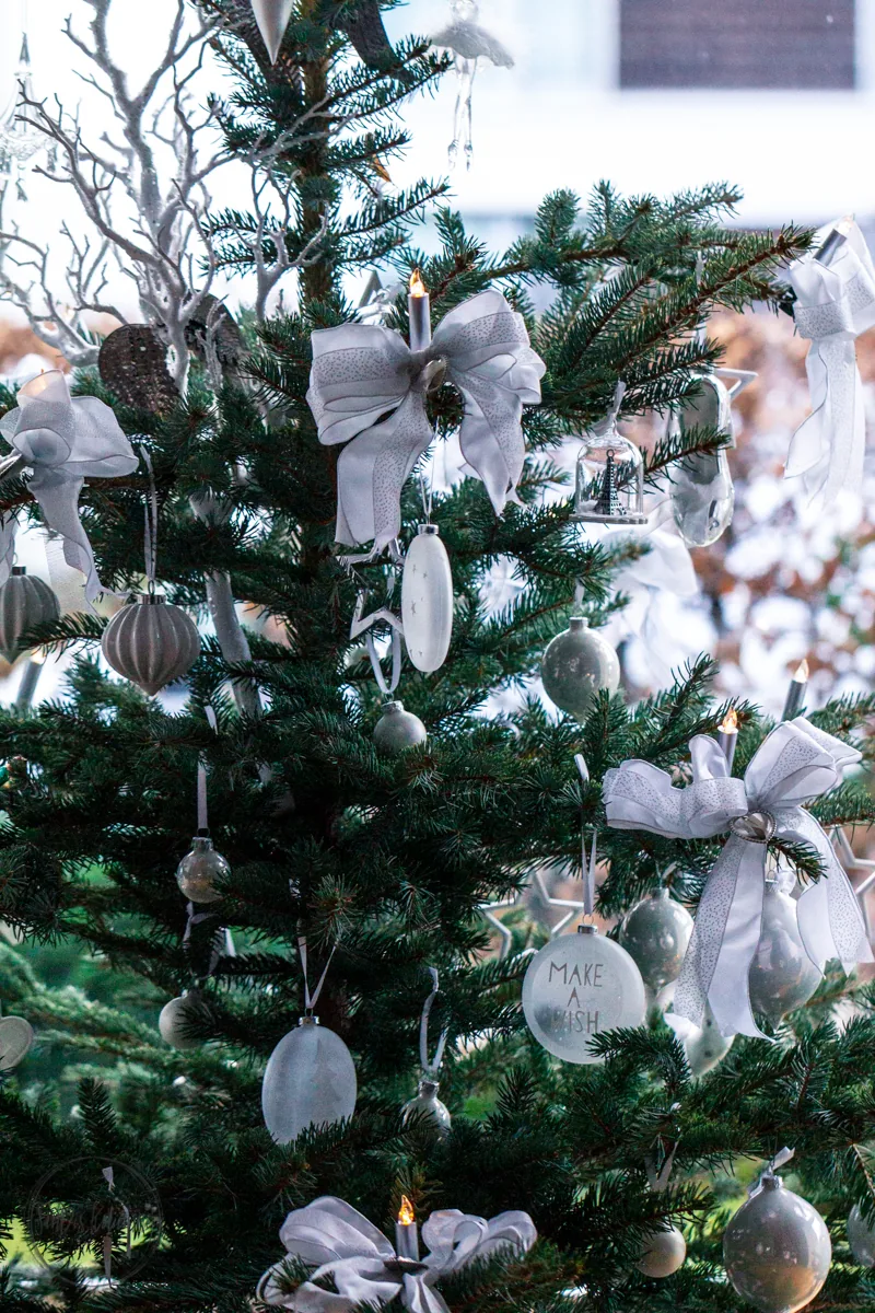 Zweigpartie mit weißen Schleifen, matten Kugeln, brennenden Baumkerzen und einem runden Anhänger mit der Aufschrift Make a Wish