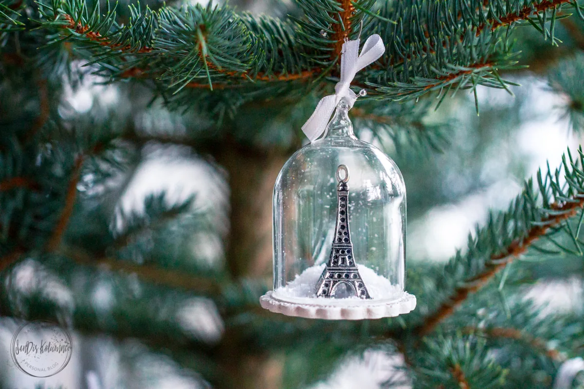 Kleine Glasglocke mit silbernem Mini-Eiffelturm auf Kunstschnee, mit weißer Schleife an einen Tannenzweig gebunden