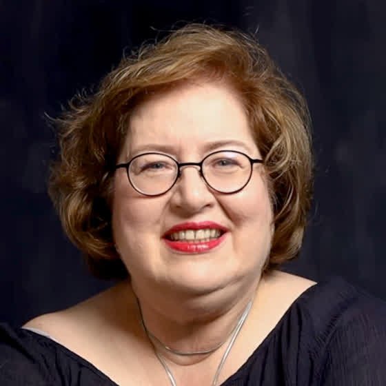 Sandra heute, in der Sandrinscher-Bluse vor der dunkelblauen Studiowand