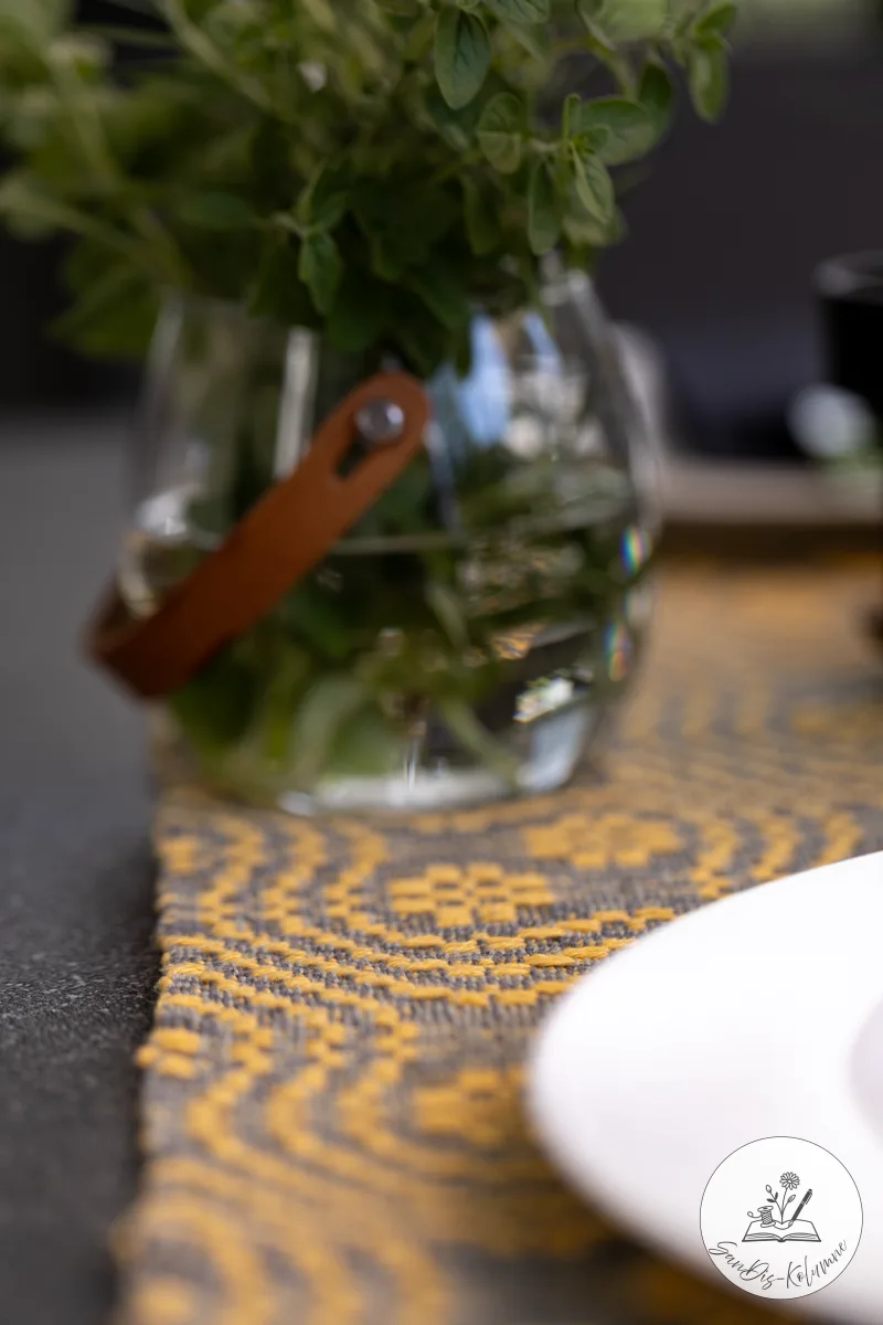 Glas mit Lederhenkel und frischem Kräuterstrauß auf dem gelb-grau gewebten Läufer, rechts der Rand eines weißen Tellers