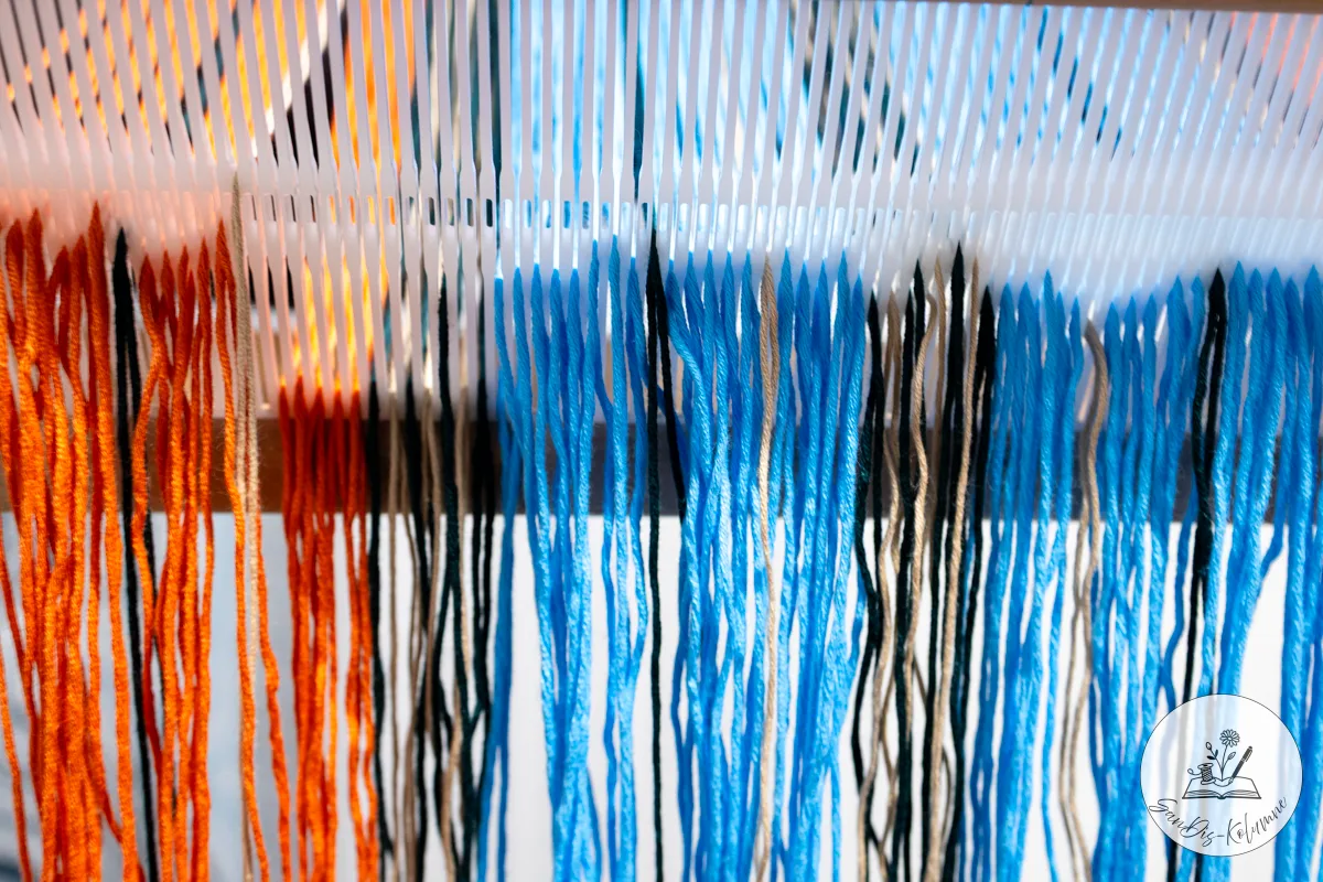 Nahaufnahme des weißen Gatterkamms, durch dessen Schlitze bunte Kettfäden gezogen sind und locker herabhängen