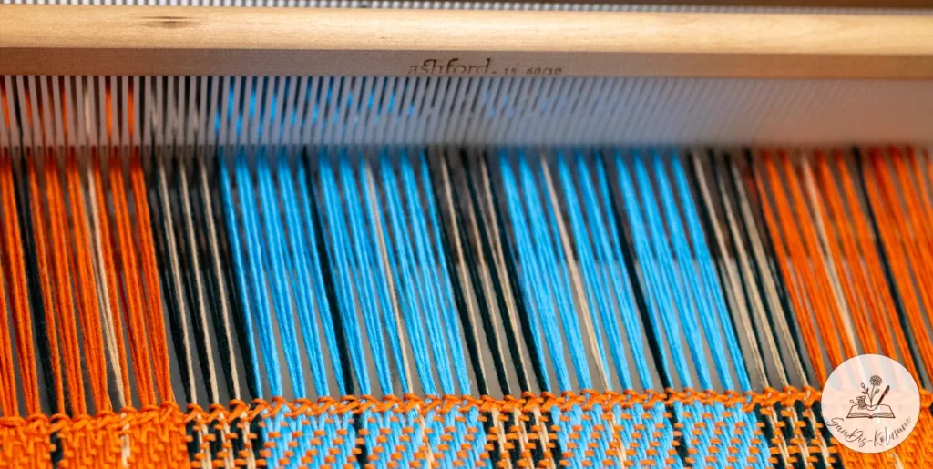 Kettfäden in Orange, Himmelblau, Dunkelgrün und Beige laufen durch den Ashford-Webkamm, darunter beginnt das Karogewebe