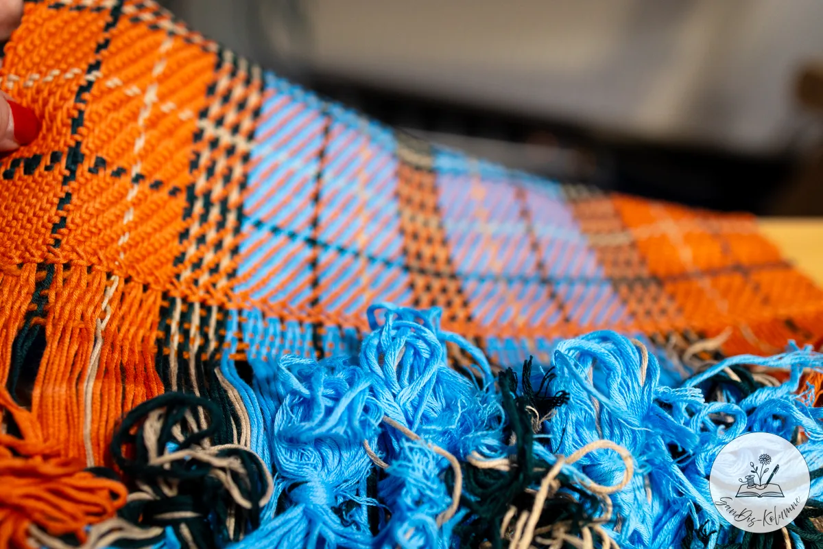 Unter der angehobenen Ecke des orange-blauen Karogewebes wartet ein Gewirr offener Fransen in Hellblau, Dunkelgrün und Beige