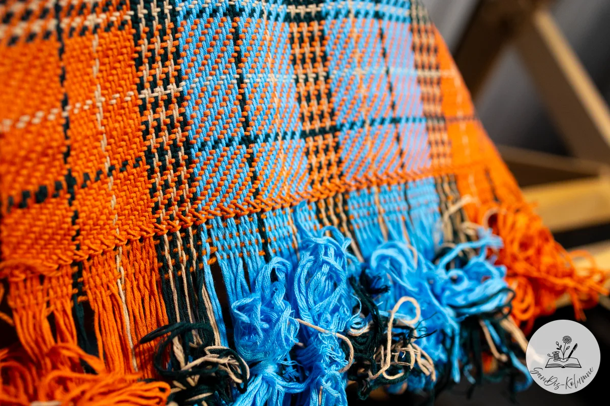 Das Karogewebe in Orange und Himmelblau liegt über einer Kante, darunter hängen die noch unversäuberten Fransen kringelig herab