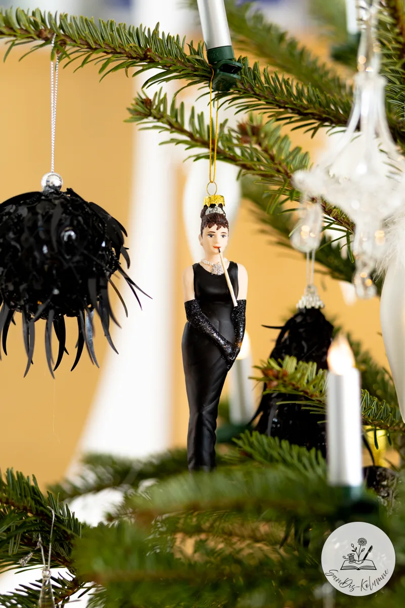 Christbaumanhänger als Audrey-Hepburn-Figur im schwarzen Kleid mit Zigarettenspitze, daneben eine schwarze Federkugel