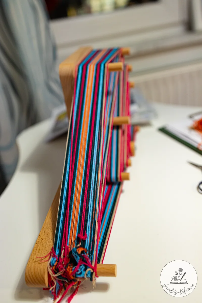 Dicht gewickelte Kette in Pink, Orange, Hellblau und Dunkelgrün auf dem hochkant stehenden Webrahmen, unten geknotete Fransen