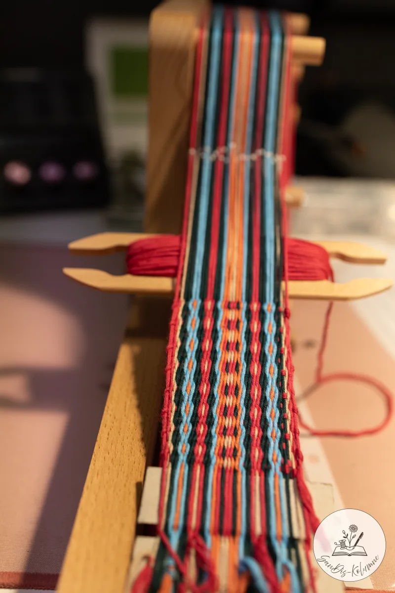 Blick entlang des halb fertigen Webbandes mit rot-blauem Punktmuster, das Schiffchen mit rotem Schussfaden liegt bereit
