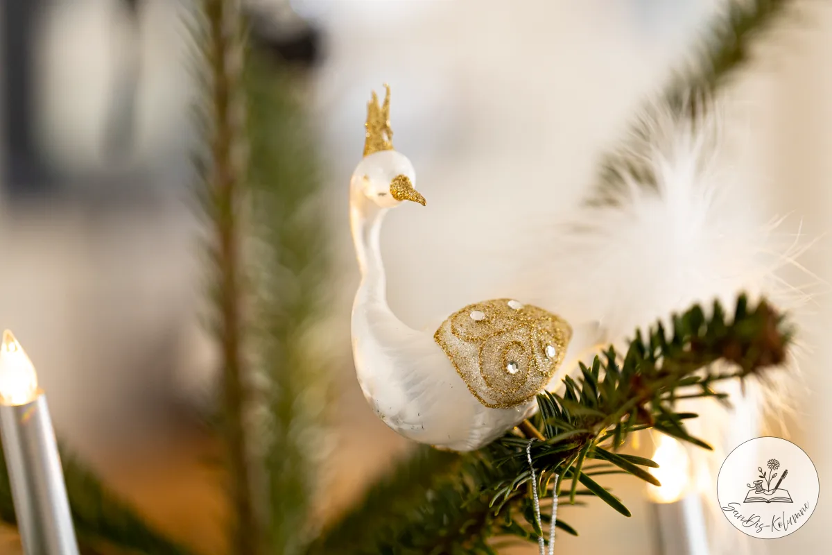 Weißer Glasvogel mit goldenem Krönchen und flauschigem Federschwanz, angeklammert an einen Tannenzweig