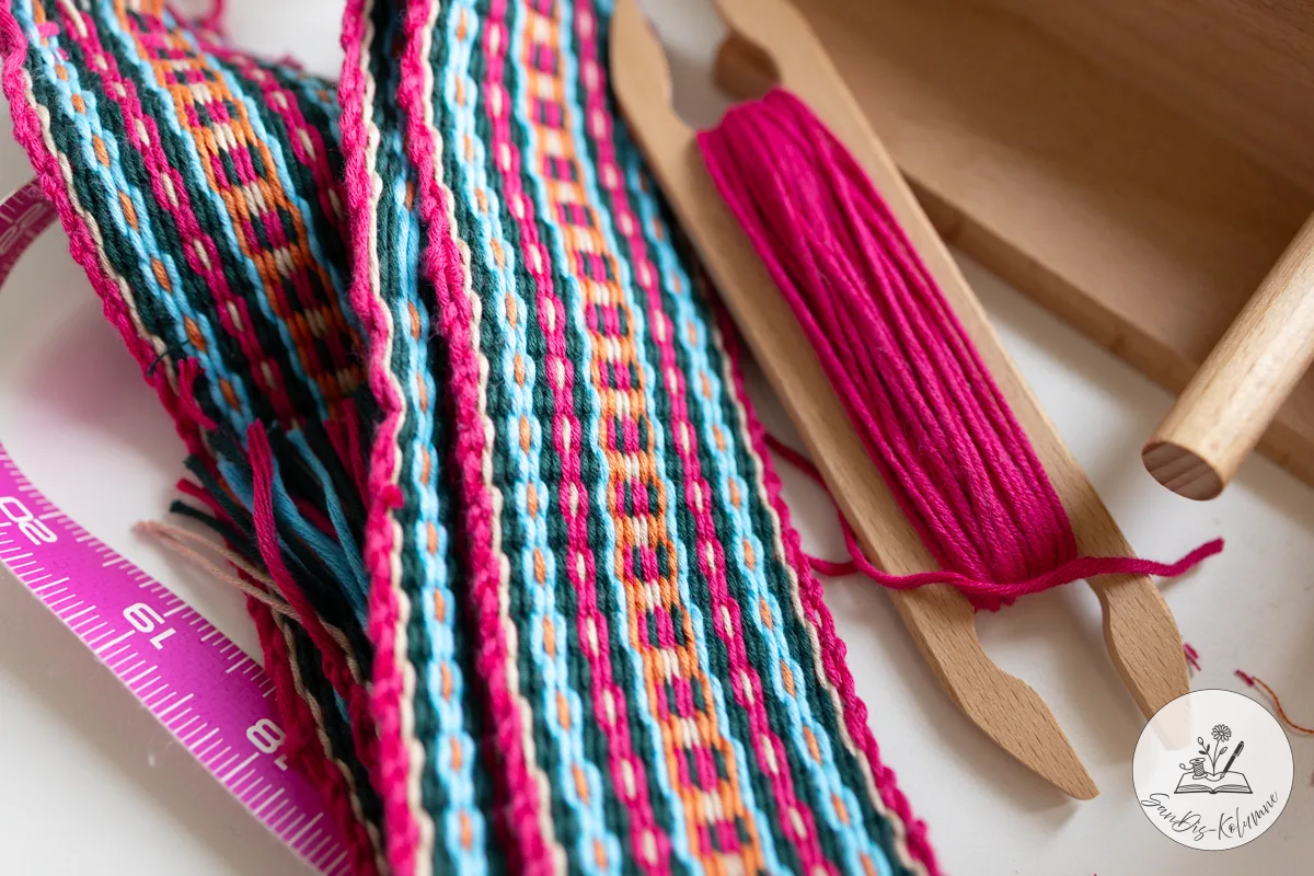Nahaufnahme des gewebten Bandes in Pink und Türkis, rechts ein Webschiffchen aus Holz mit pinkem Garn umwickelt