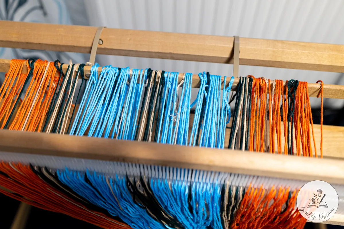Kettfäden in Orange, Hellblau, Petrol und Beige laufen über die Holzleiste eines Webrahmens zum Gatterkamm