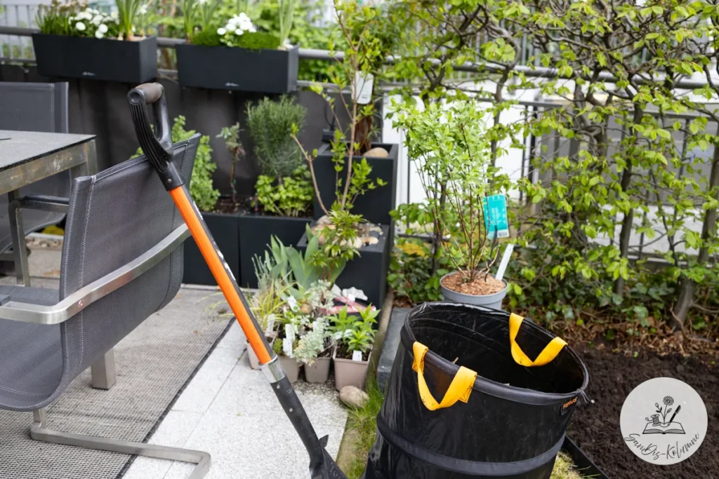Orange-schwarzer Spaten lehnt am Gartenstuhl, daneben ein Pop-up-Gartensack mit gelben Griffen und junge Stauden in kleinen Töpfen
