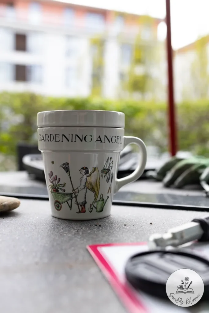 Weiße Tasse in Blumentopf-Form mit Deckel, Aufschrift Gardening Angel und gezeichnetem Engel mit Schubkarre und Rechen auf dem Gartentisch