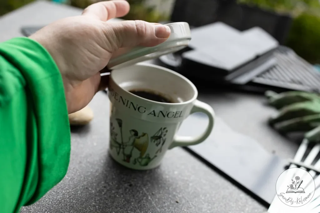 Eine Hand im grünen Pullover hebt den Deckel von der Gardening-Angel-Tasse mit schwarzem Kaffee auf dem grauen Gartentisch