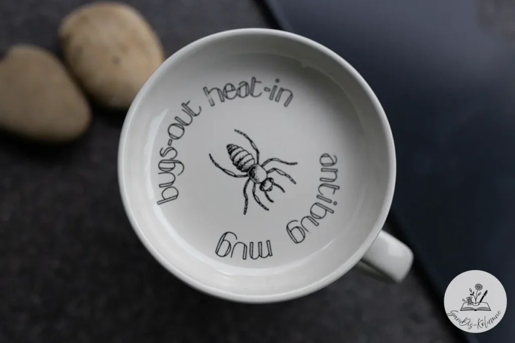 Weiße Tasse von oben: gezeichnete Ameise und der runde Schriftzug bugs-out heat-in antibug mug, daneben zwei helle Kieselsteine
