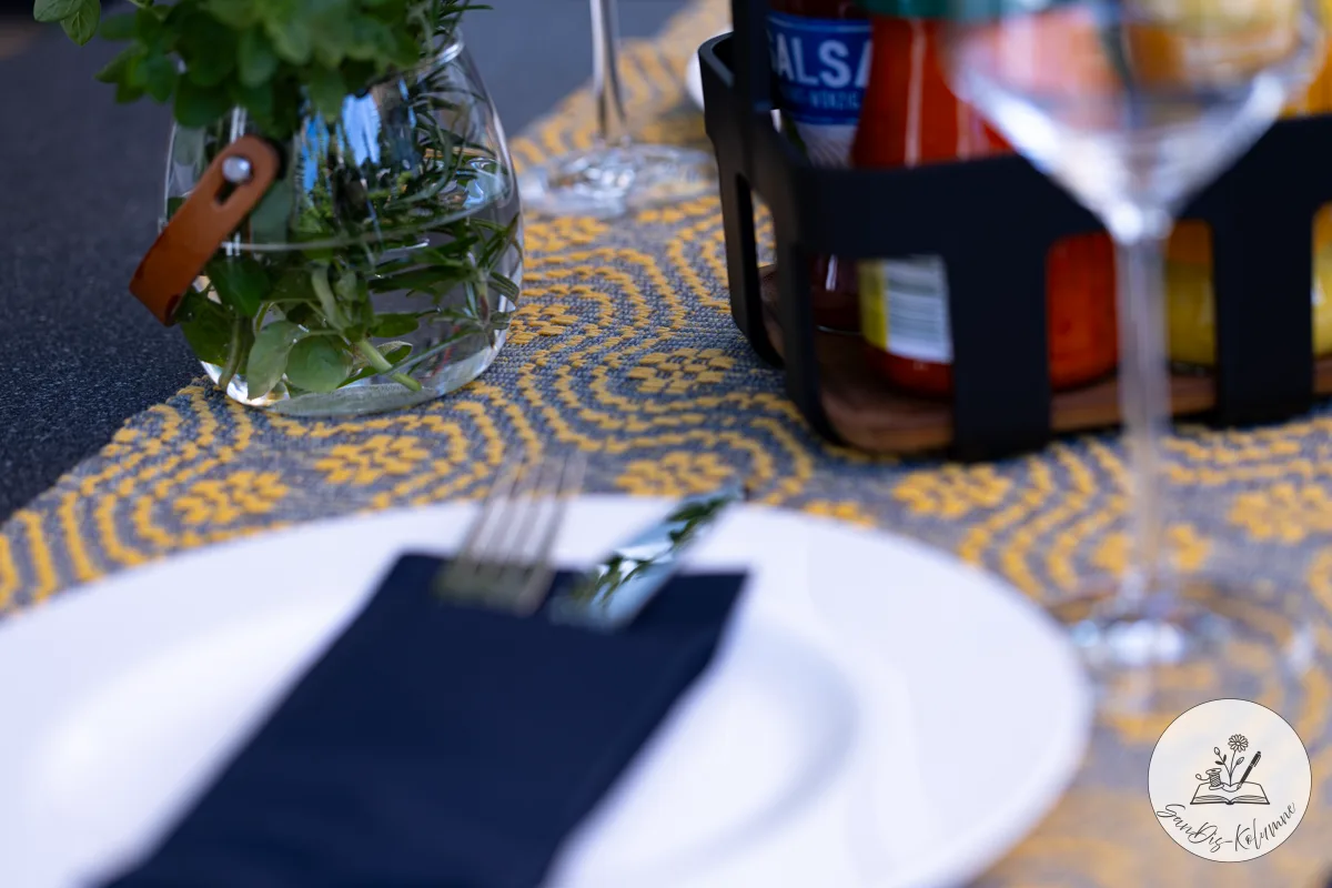 Weißer Teller mit dunkelblauer Serviette und Gabel auf dem gelb-grau gemusterten Tischläufer, dahinter Kräuterglas und ein Salsa-Glas