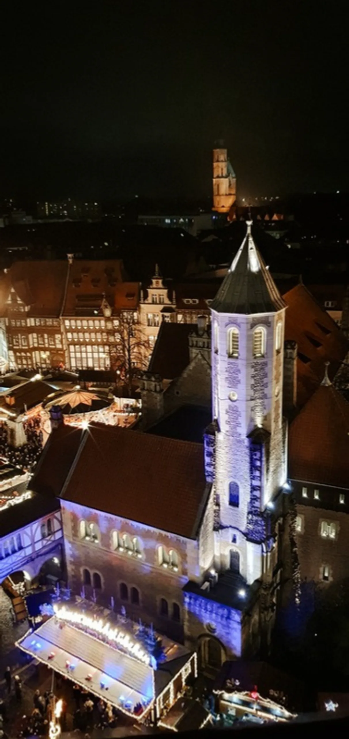 Blick vom Rathausturm auf einen angestrahlten Kirchturm über dem nächtlichen Weihnachtsmarkt, am Fuß leuchten die Dächer der Marktstände