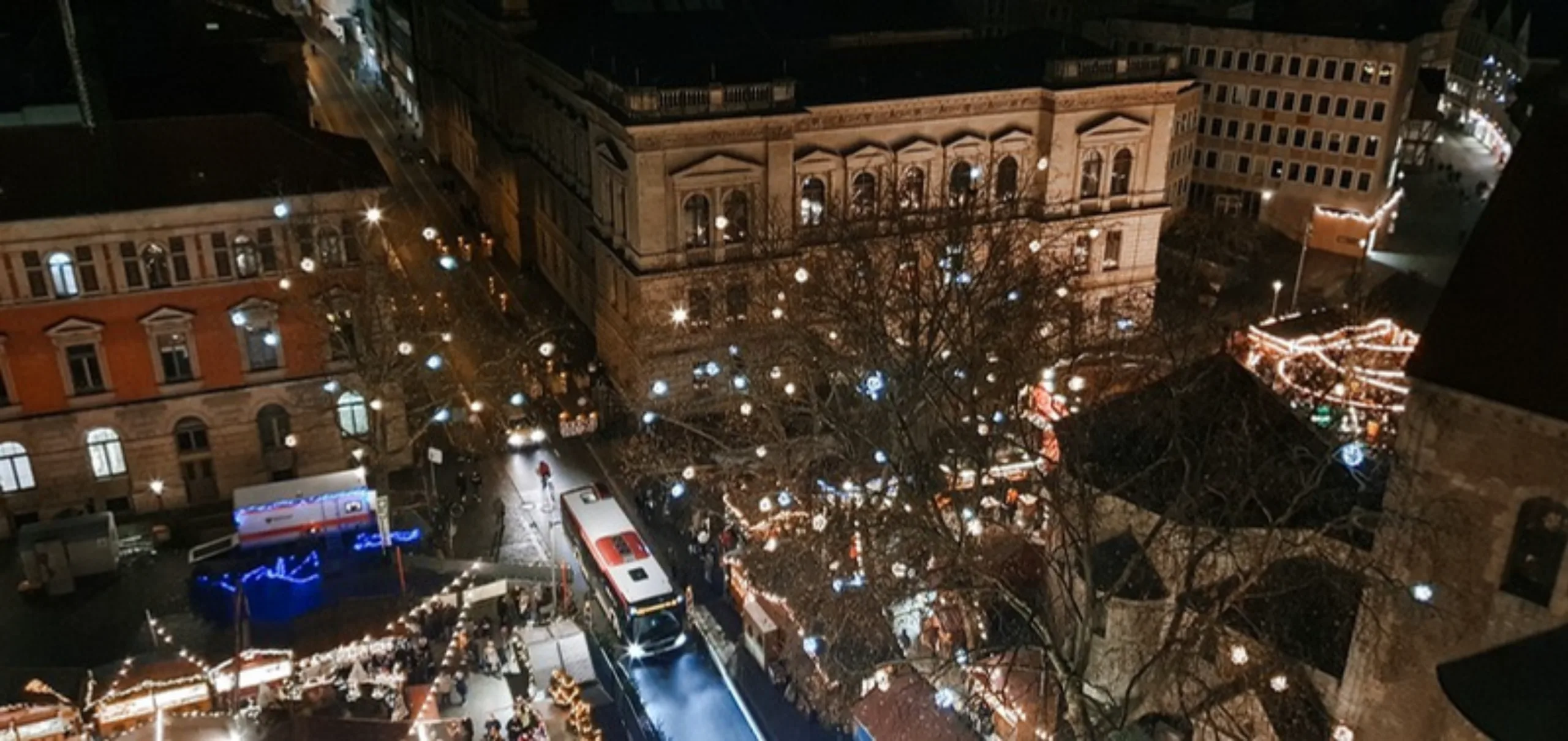 Blick vom Rathausturm auf die beleuchteten Buden des Weihnachtsmarkts zwischen historischen Gebäuden, durch die Straße fährt ein Bus