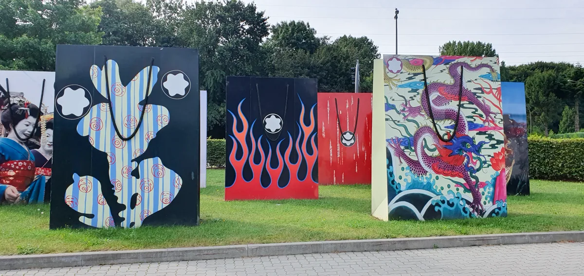 Fünf große Taschen-Skulpturen mit Kunstmotiven auf dem Museumsrasen, darunter ein lilafarbener Drache über Wellen und rote Flammen.