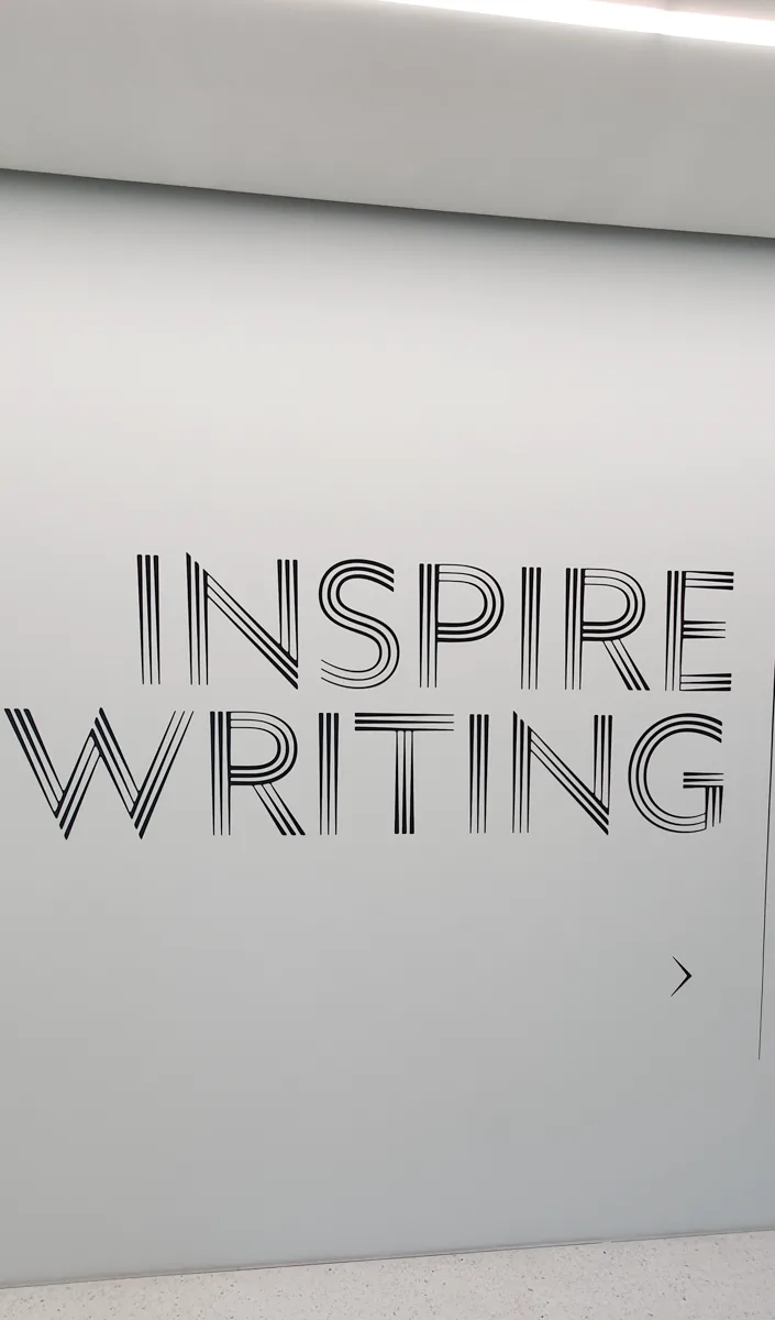 Großer Schriftzug Inspire Writing in gestreifter Outline-Typografie auf der geschwungenen hellgrauen Museumswand.