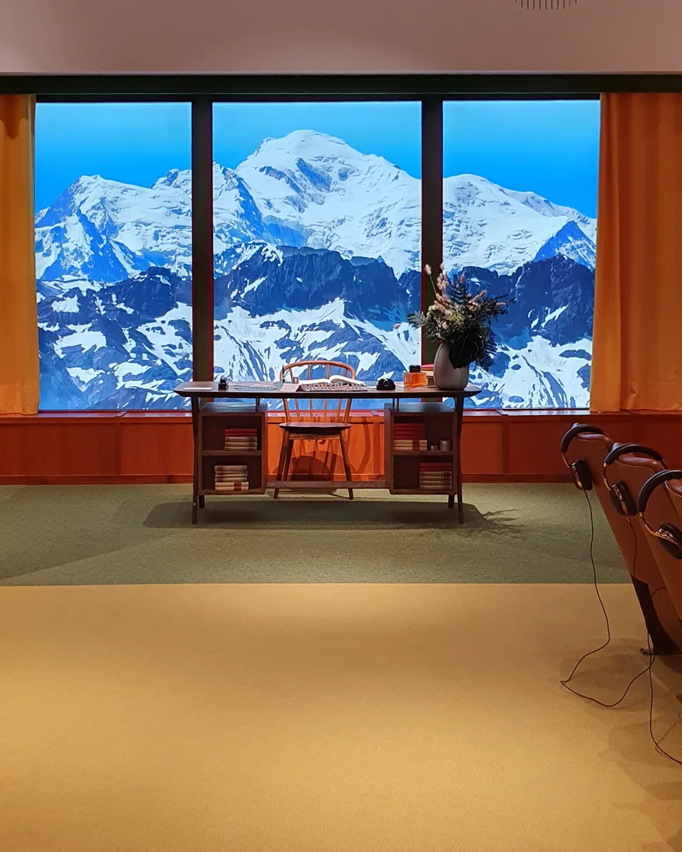 Nachgebauter Schreibtisch aus dem Wes-Anderson-Filmset vor der Panoramawand mit schneebedeckten Gipfeln, gerahmt von orangefarbenen Vorhängen.