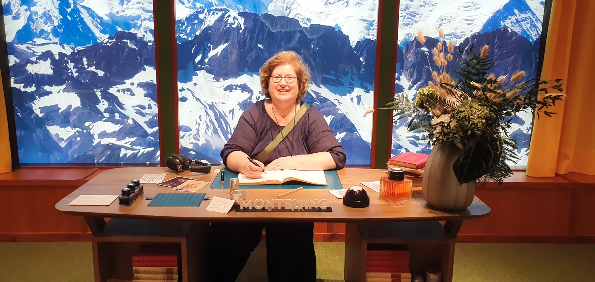 Noch einmal der Filmset-Schreibtisch vor der Bergkulisse: Sandra Dirks schreibt im warmen Licht, vorn liegen rote Bücher und dunkle Metalldosen.
