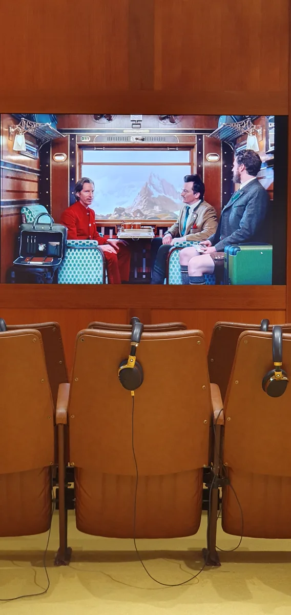 Auf dem Museumsbildschirm läuft die Zugabteil-Szene aus dem Montblanc-Kurzfilm von Wes Anderson, davor leere Ledersessel mit Kopfhörern.