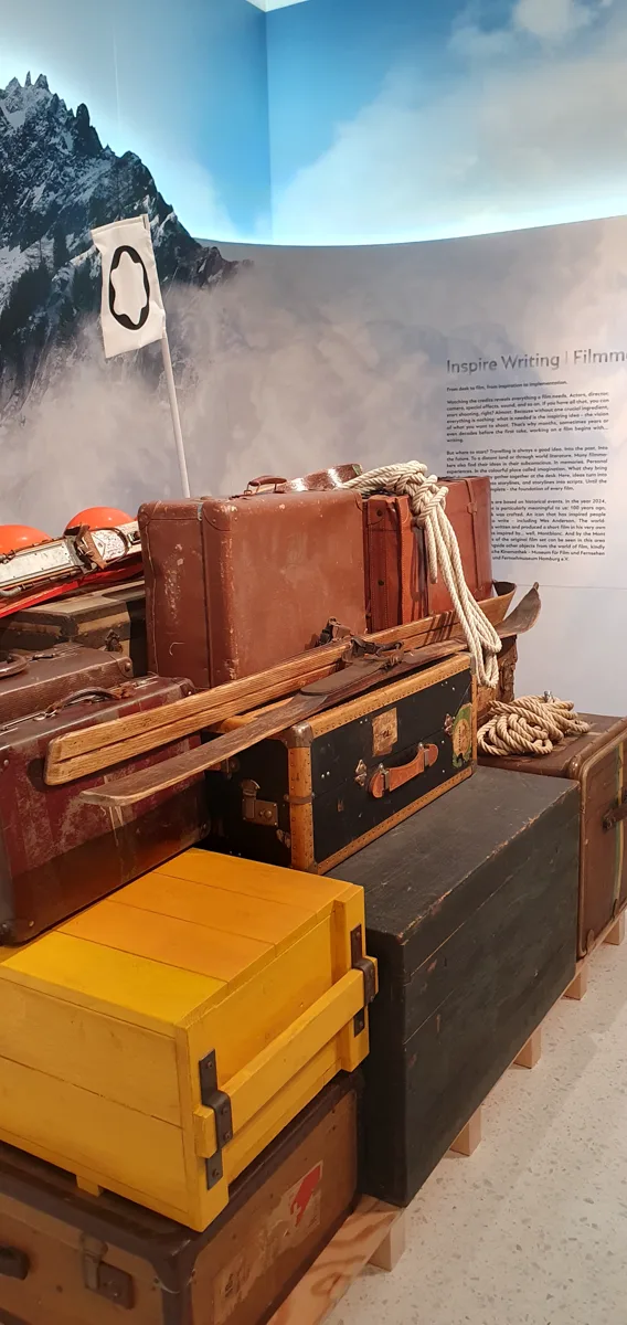 Gestapelte alte Lederkoffer, Holzski und Tauwerk vor der Bergpanorama-Tapete, obenauf eine weiße Fahne mit dem Montblanc-Stern, rechts die Ausstellungstafel.