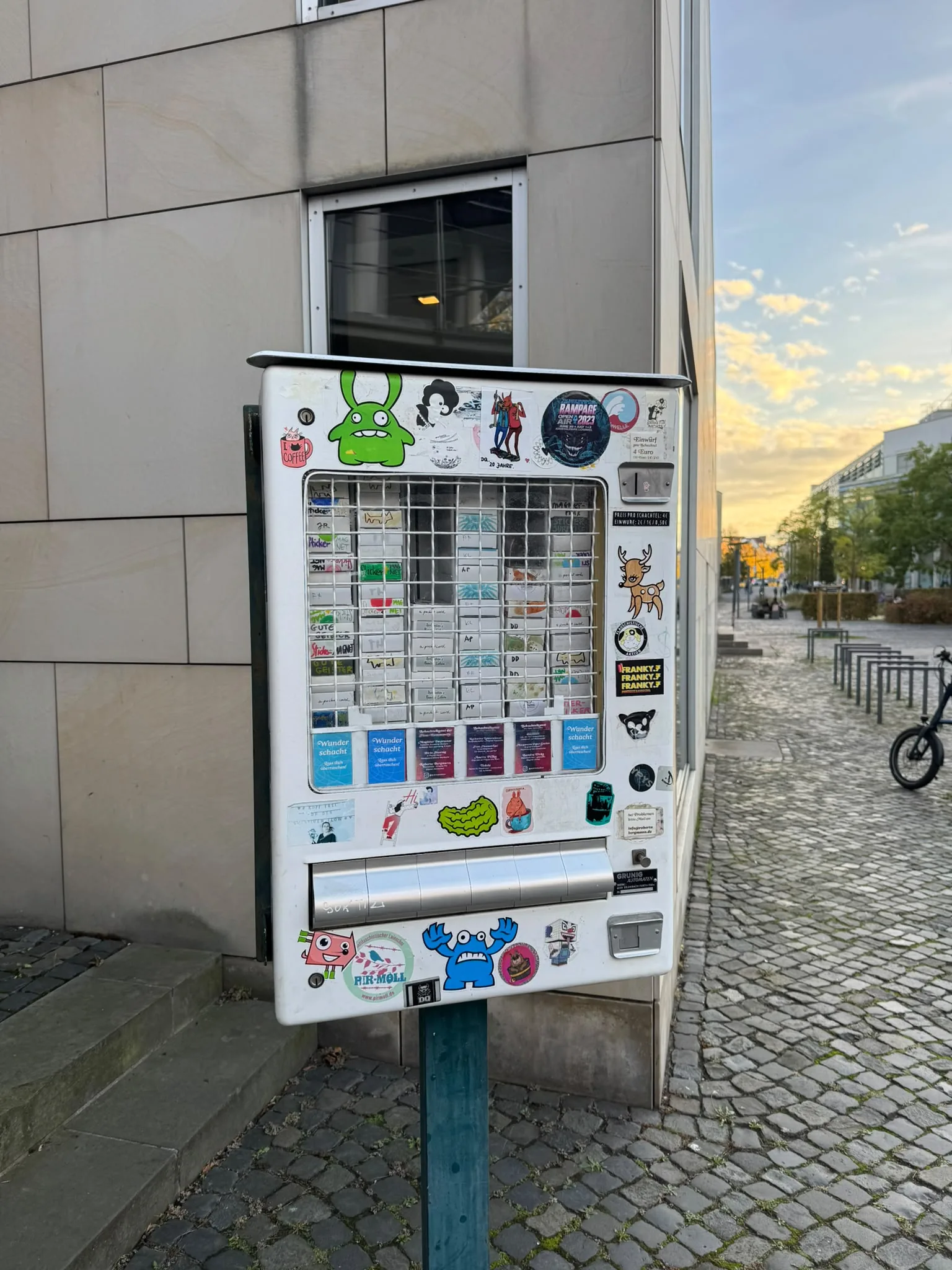 Der Kunstautomat, ein ehemaliger Zigarettenautomat voller Aufkleber, steht auf einem Pfahl vor dem Kleinen Haus des Staatstheaters Braunschweig, daneben Kopfsteinpflaster und Abendhimmel.