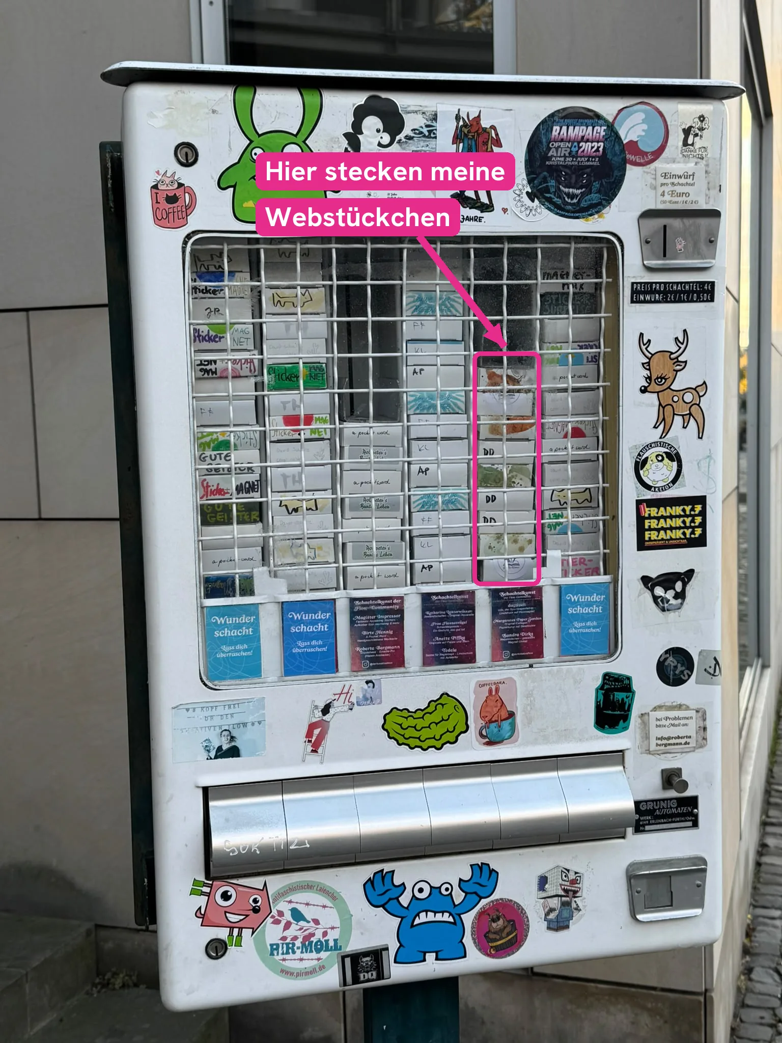 Frontansicht des Kunstautomaten in Braunschweig, ein pinker Rahmen markiert die fünfte Schachtelreihe von links, in der die Webstückchen von Sandra Dirks stecken.