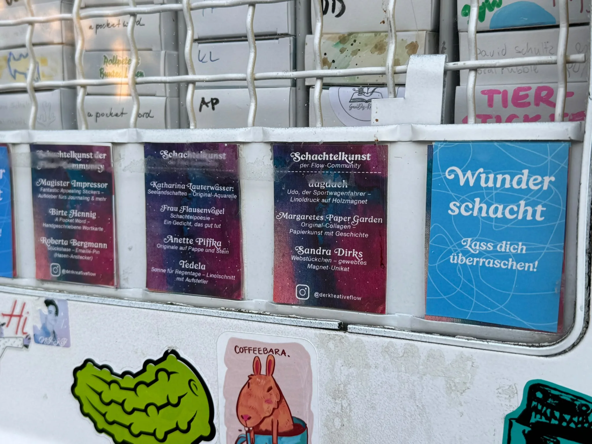 Hinter dem Gitter des Kunstautomaten listen Schilder die Schachtelkunst der Flow-Community auf, darunter Sandra Dirks mit Webstückchen, gewebtes Magnet-Unikat.